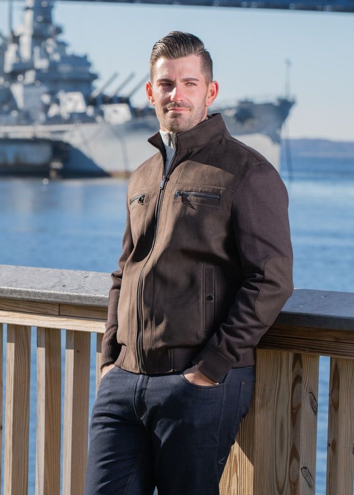 Man leaning on a wooden railing by the water, wearing a brown jacket with a naval ship in the background.