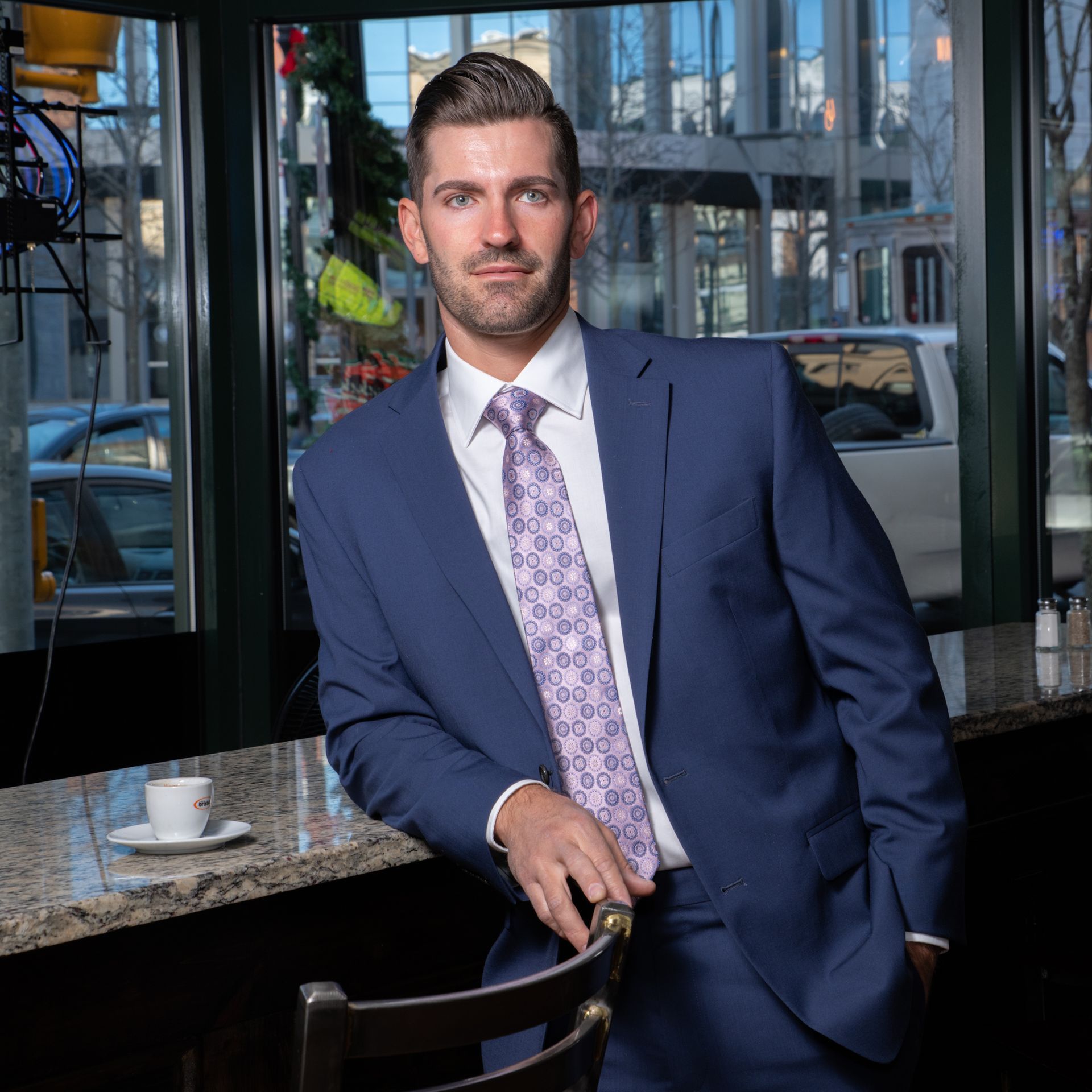 Man in a blue suit leaning at a café counter with a coffee cup nearby