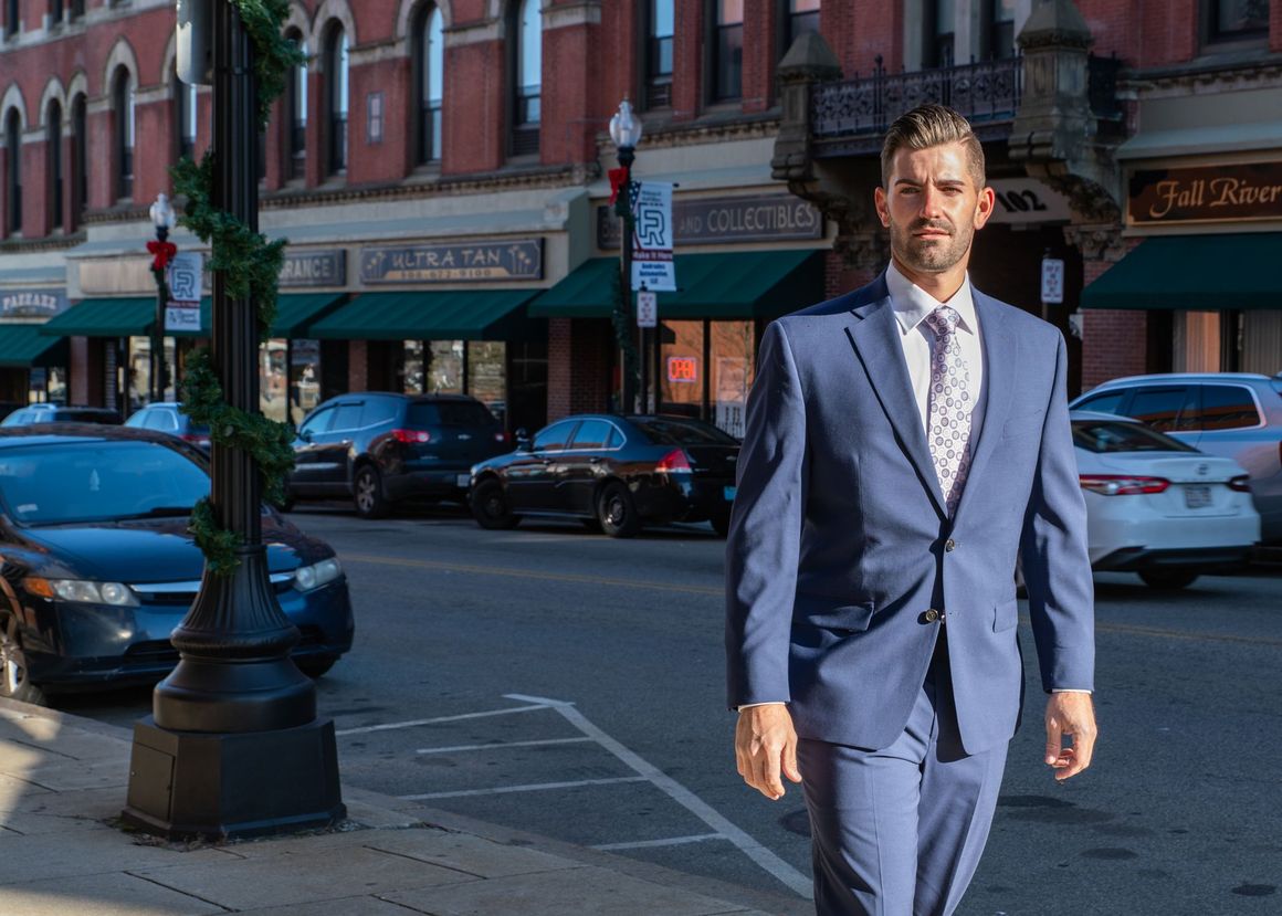 Man in a blue suit walking on a city street with parked cars and storefronts behind him