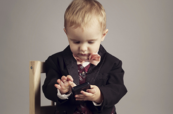 Young child in a black suit looking down at a smartphone, with a wooden chair beside them
