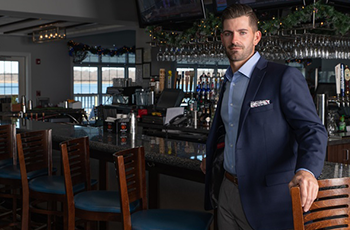Man in navy blazer standing in a dimly lit bar, leaning on a chair beside the counter.