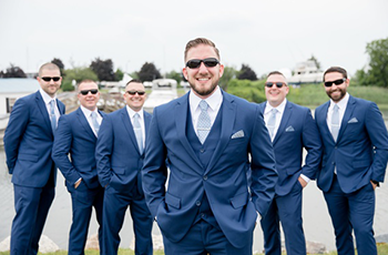 Six men in blue suits and sunglasses standing outdoors, with one man centered in front smiling.