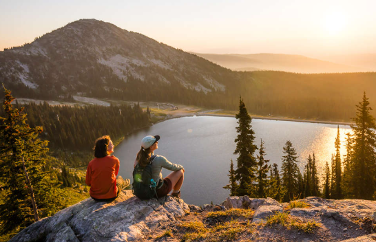 Two hikers sit on a rocky overlook above a sunlit lake and forested mountains at sunset