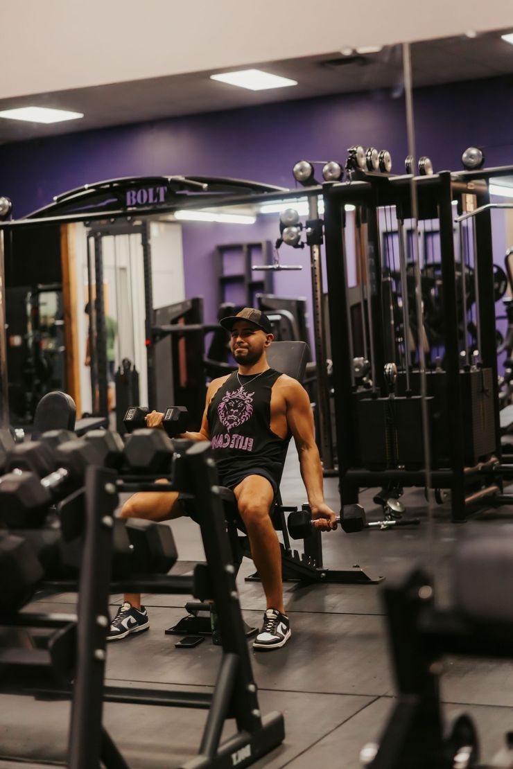 Man seated on a gym machine in a purple-lit fitness center, with dumbbells and cable equipment nearby.