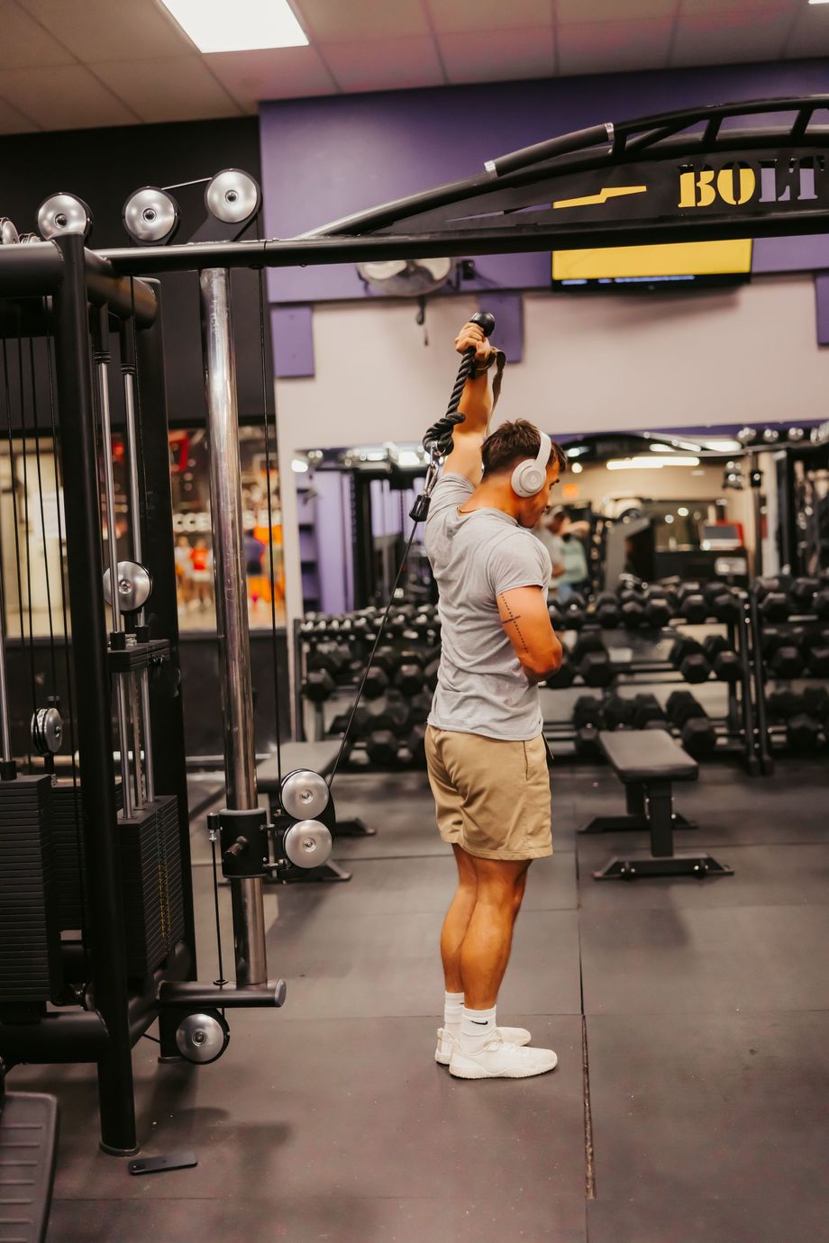 Person using a cable machine in a gym, reaching overhead with one arm.