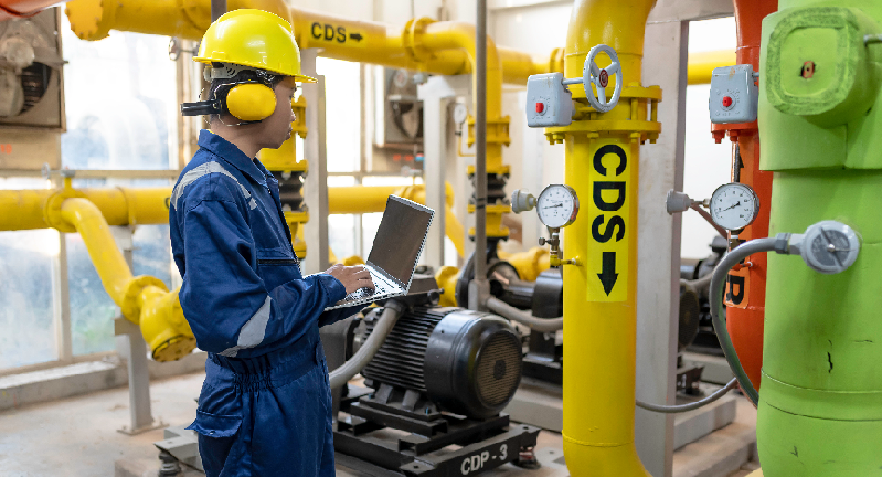 A technician in a hard hat and protective gear inspects machinery and yellow pipes labeled