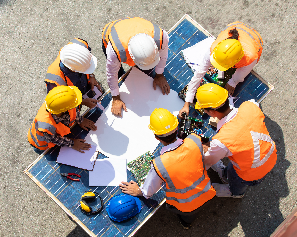 A group of construction workers in orange safety vests and hard hats reviewing blueprints on a large solar panel surface.