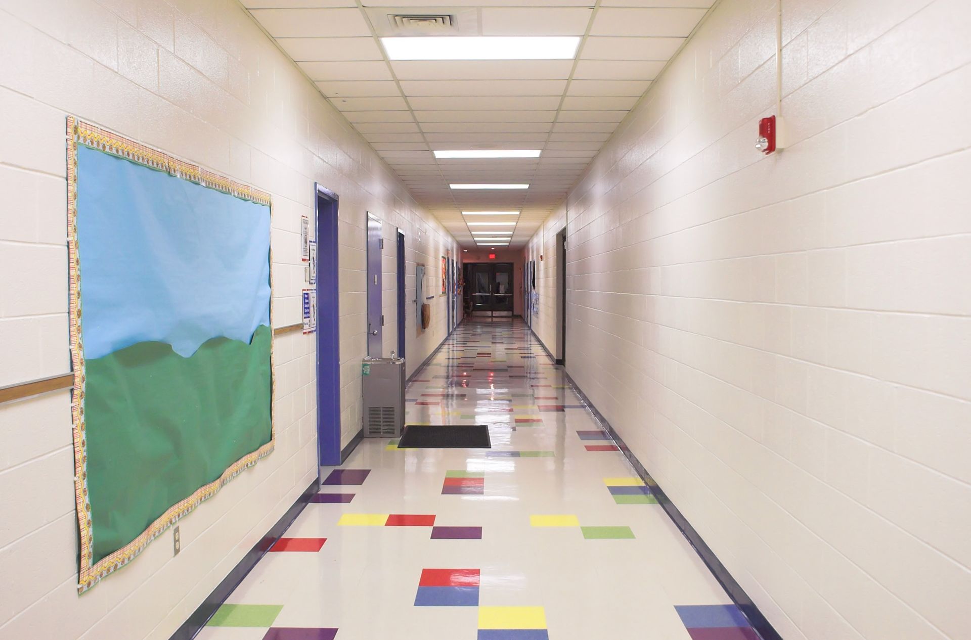 Long school hallway with tiled floor, bulletin board, and doors.