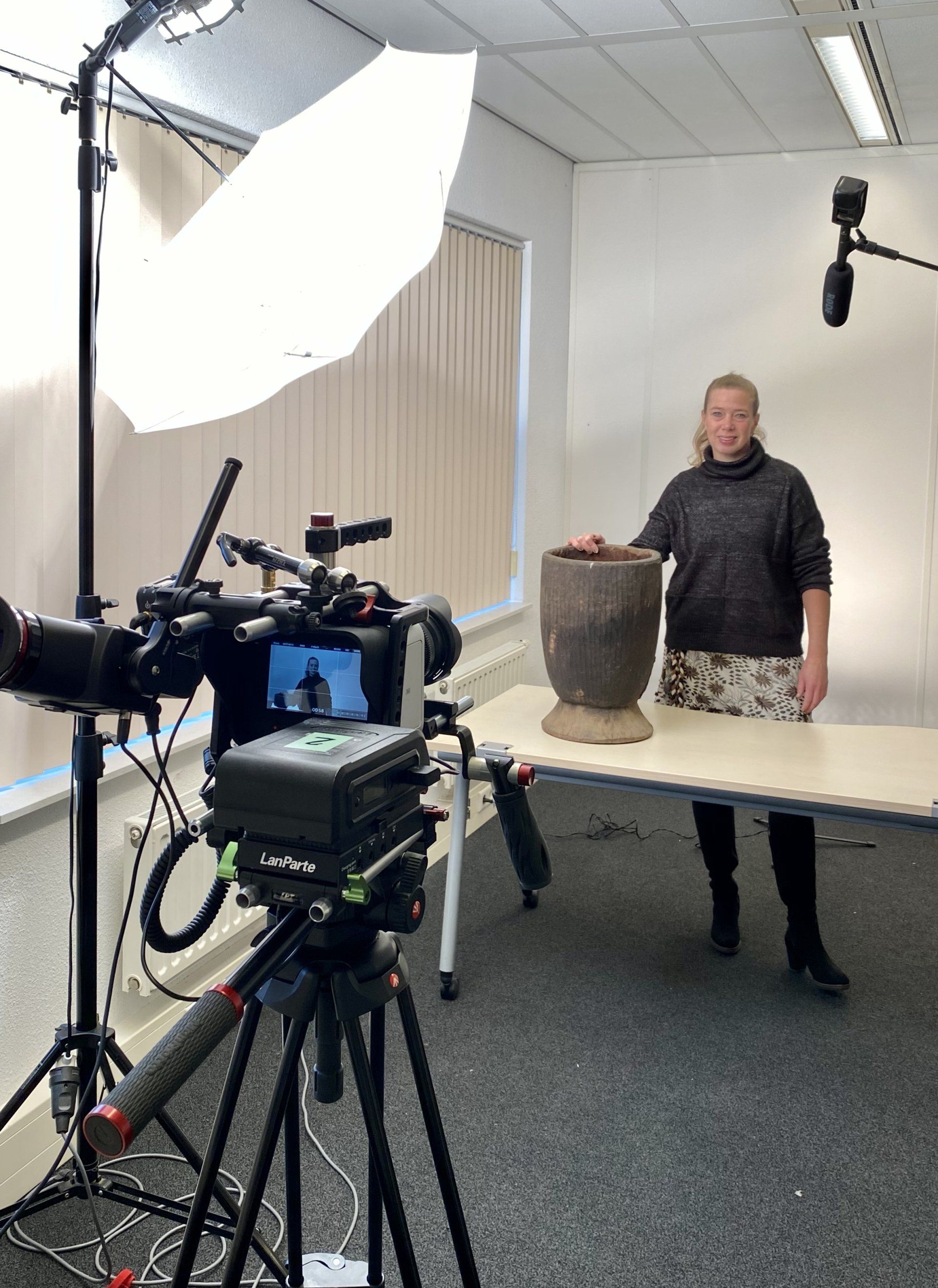 Vrouw in een studio met camera-opstelling, staat naast een houten vaas op een tafel.