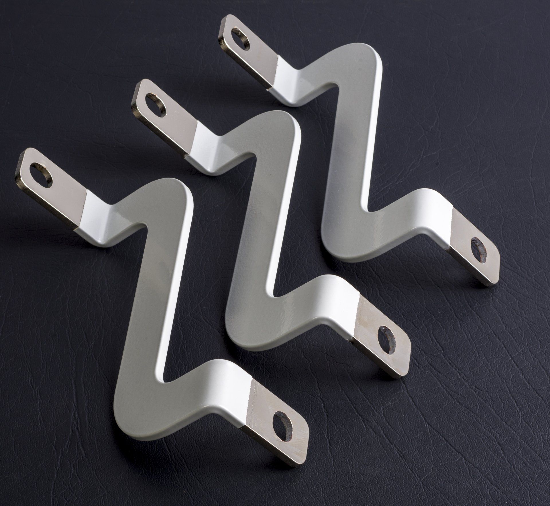 Drie zilverkleurige, Z-vormige metalen beugels met gaten aan beide uiteinden, geplaatst op een zwart oppervlak.