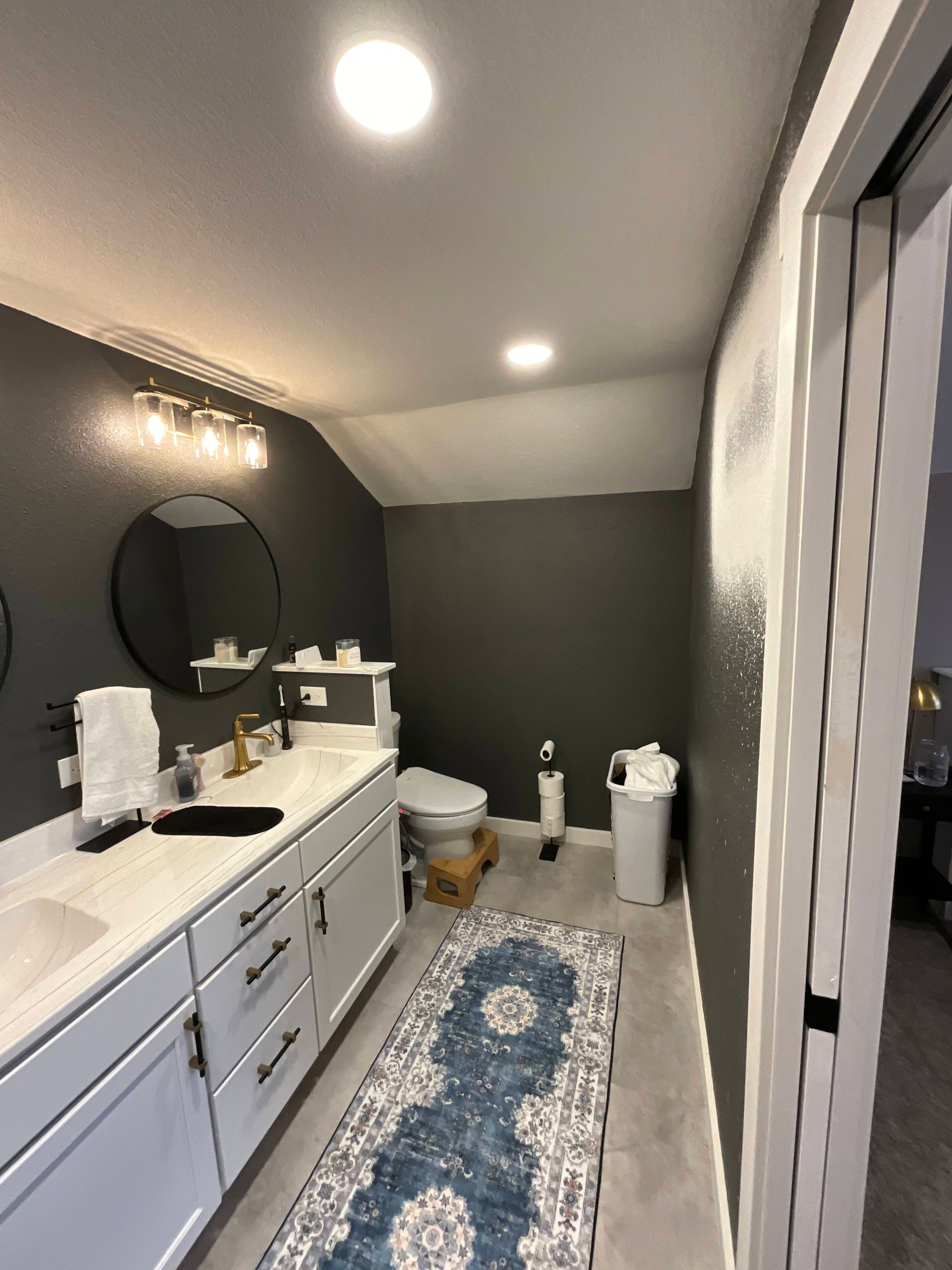 Bathroom with gray walls, white vanity, round mirror, and blue patterned rug.