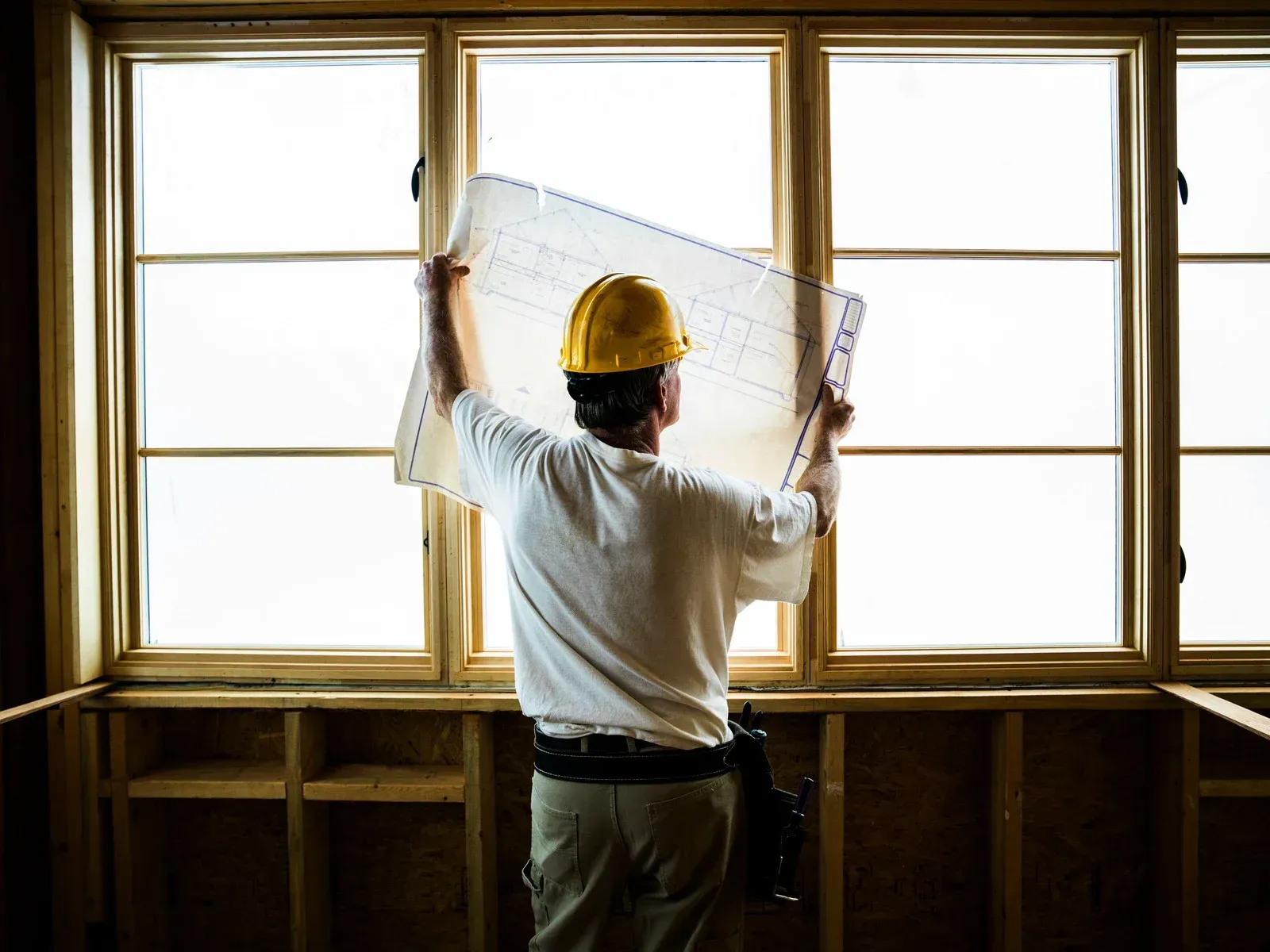 Construction worker holding blueprints in front of window, inspecting framing.