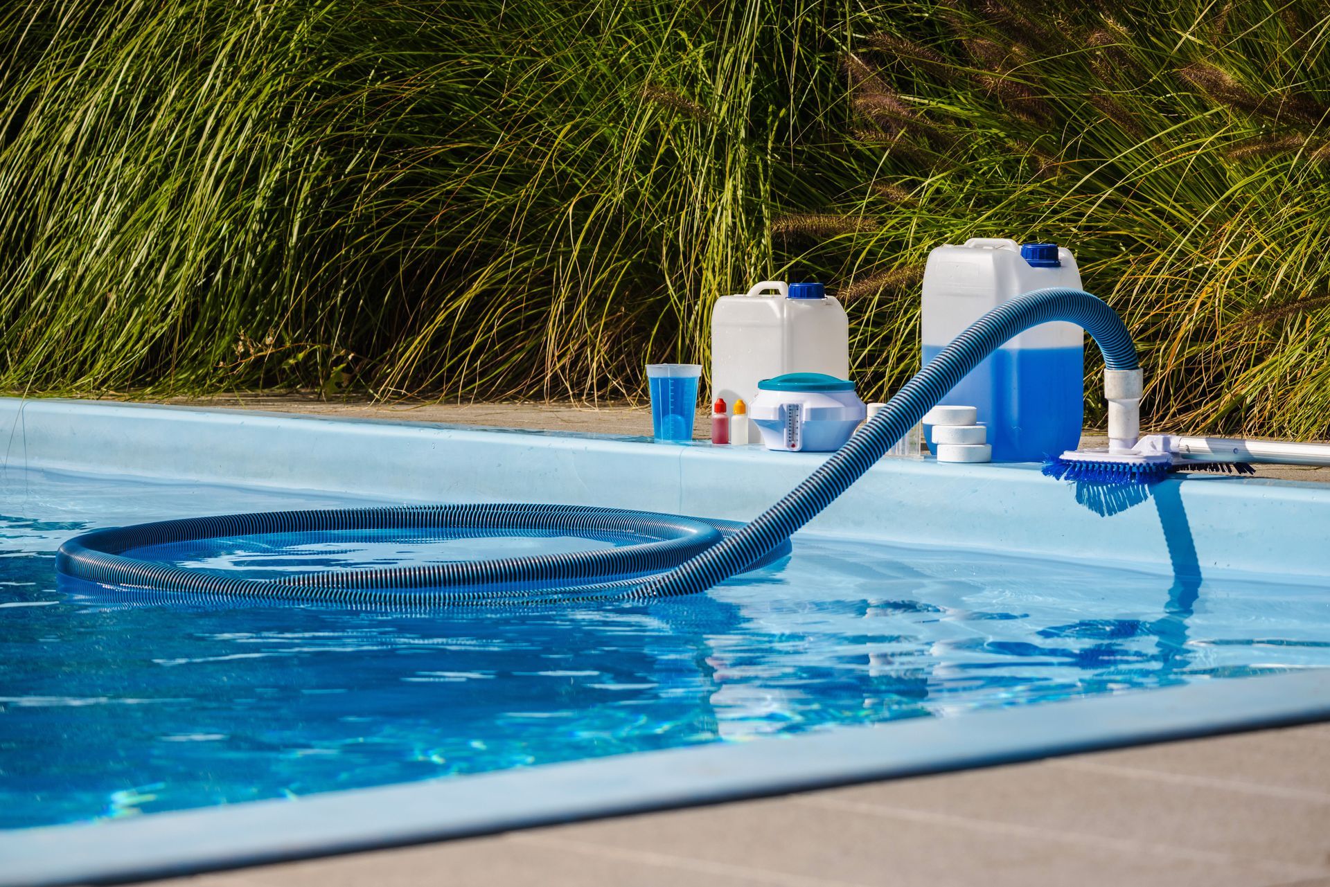 Pool vacuum hose floating in a blue swimming pool beside cleaning supplies on the deck