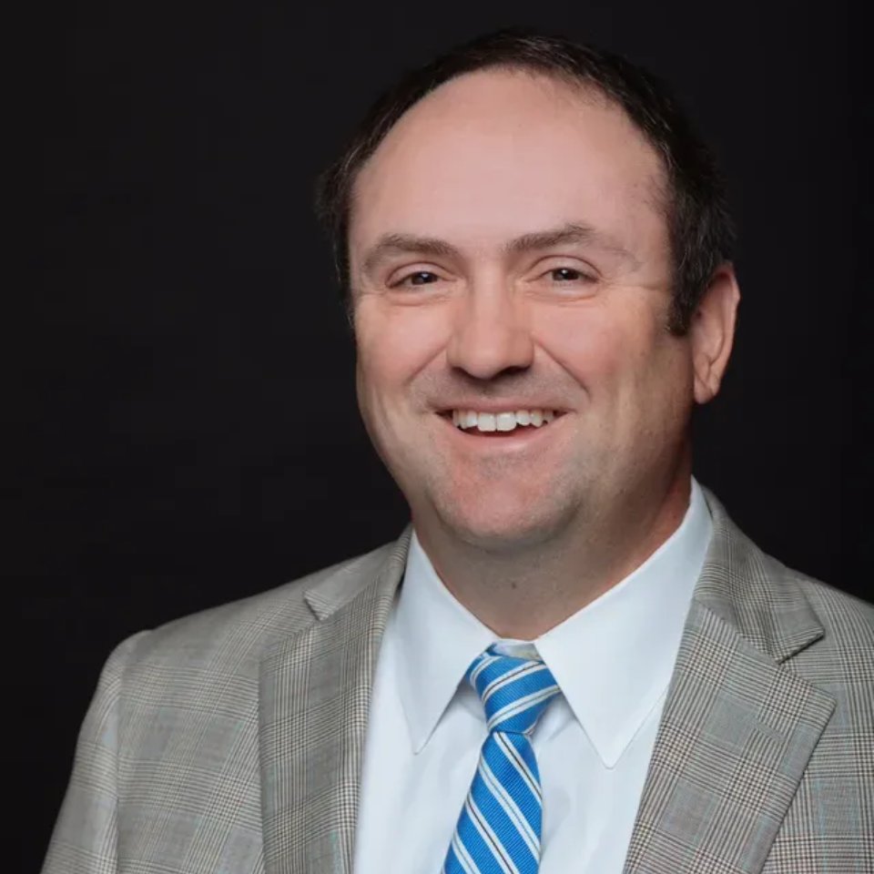 Smiling man in a light gray suit and blue striped tie against a dark background
