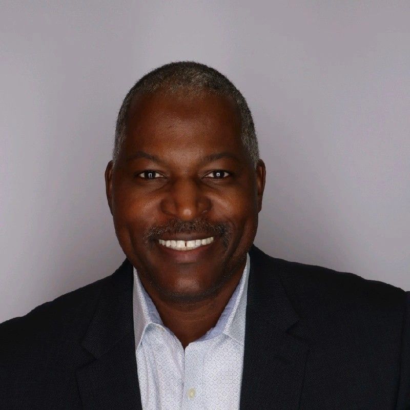 Smiling man in a dark blazer and light shirt against a plain gray background