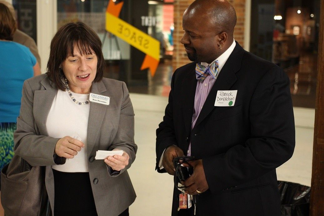 Two people conversing indoors, one gesturing while holding a small item.