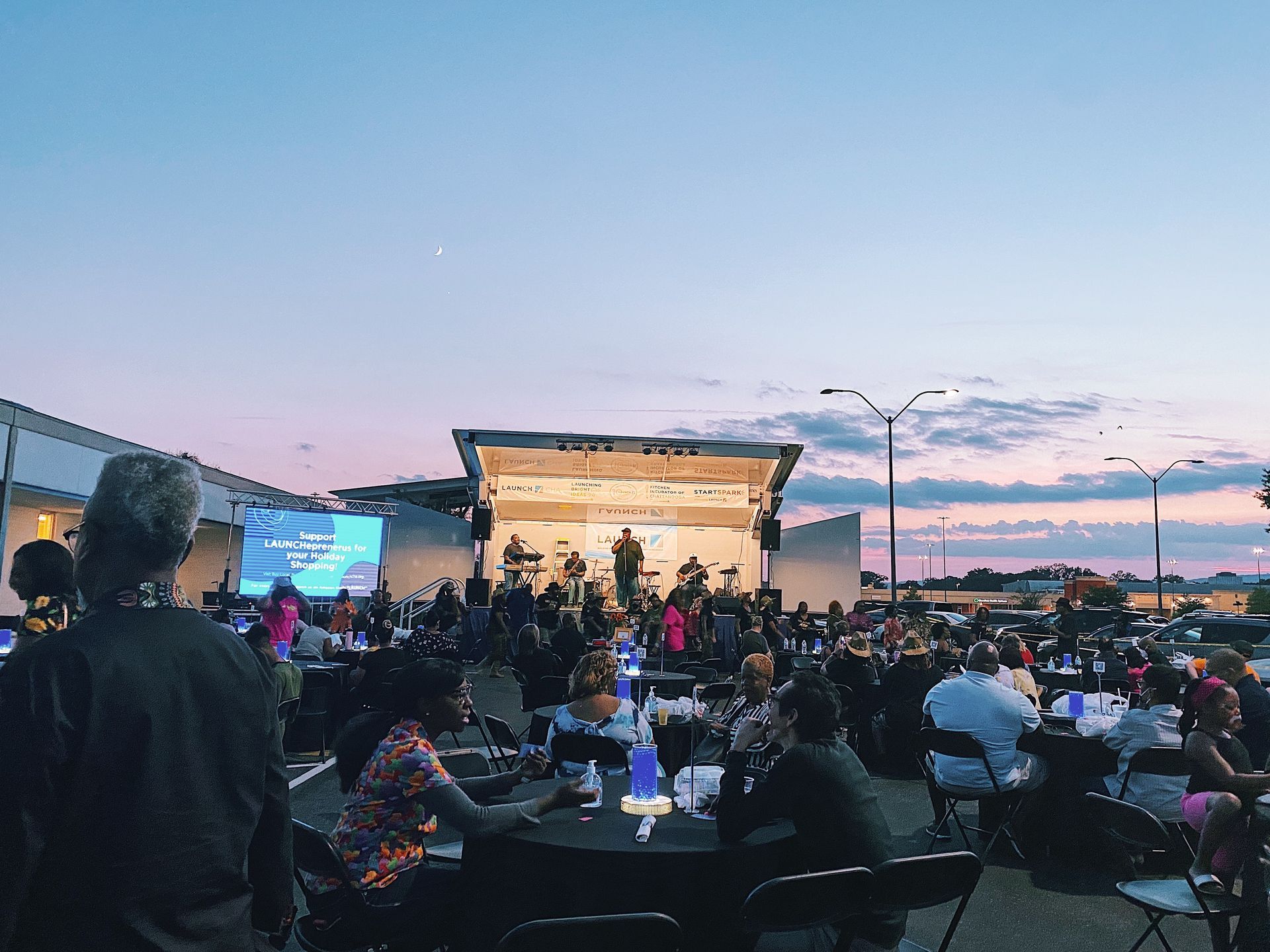 Outdoor concert at dusk with a lit stage, crowd seated at tables, and a pink-blue sunset sky.