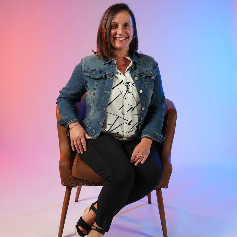 Woman seated in a brown chair, wearing a denim jacket and black pants, against a pink-blue gradient background