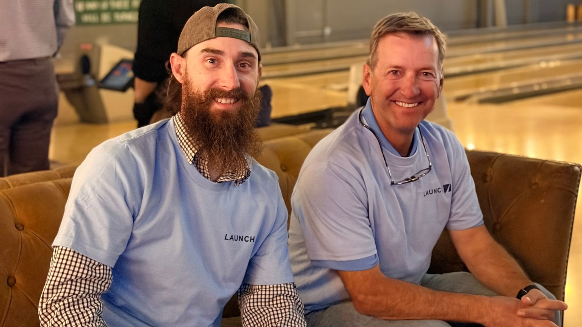 Two smiling men in light blue shirts sit on couches at a bowling alley.