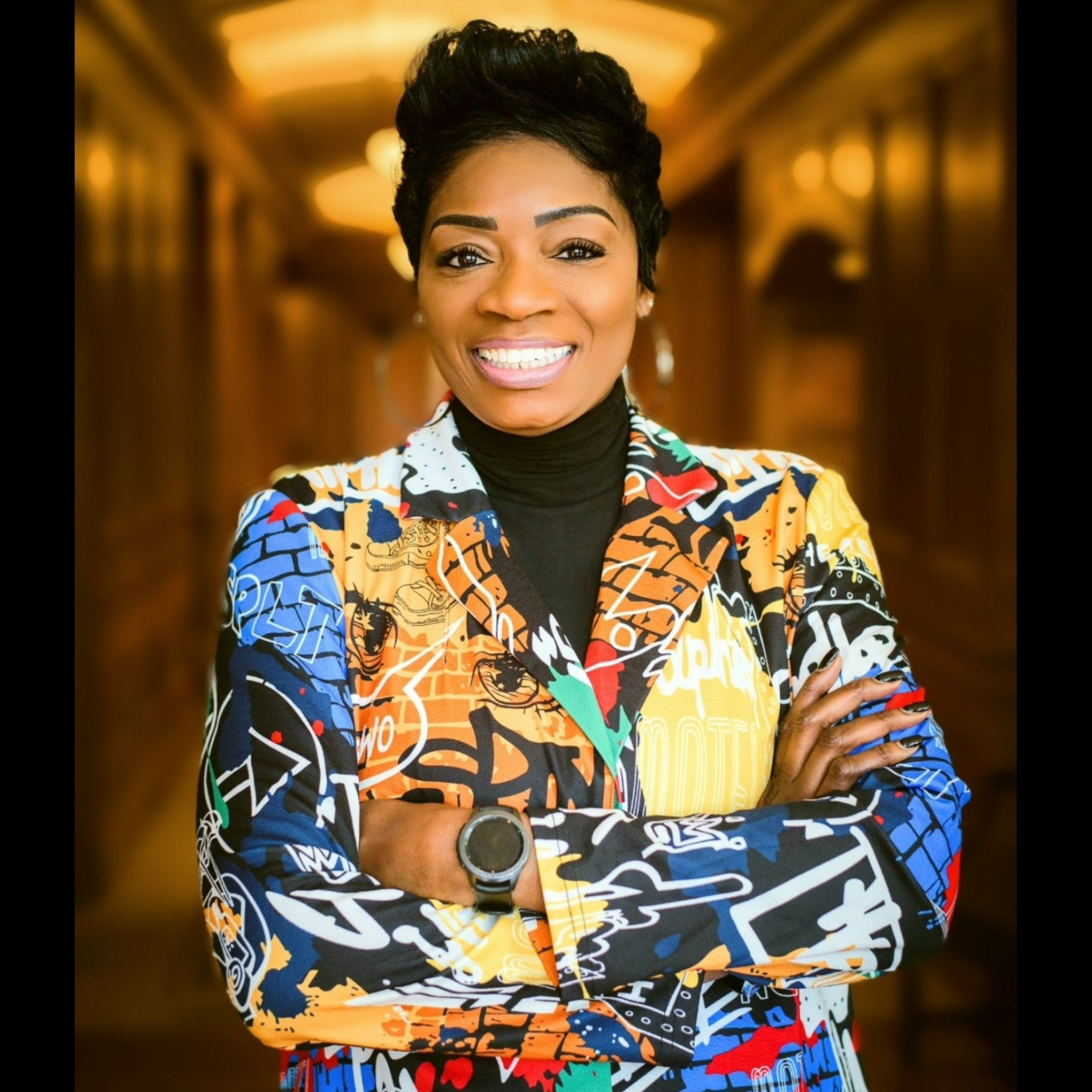 Smiling person in a colorful patterned jacket, standing with arms crossed in a warmly lit hallway.