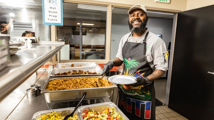 Chef serving food from a buffet line in a commercial kitchen, holding a plate and smiling at the counter