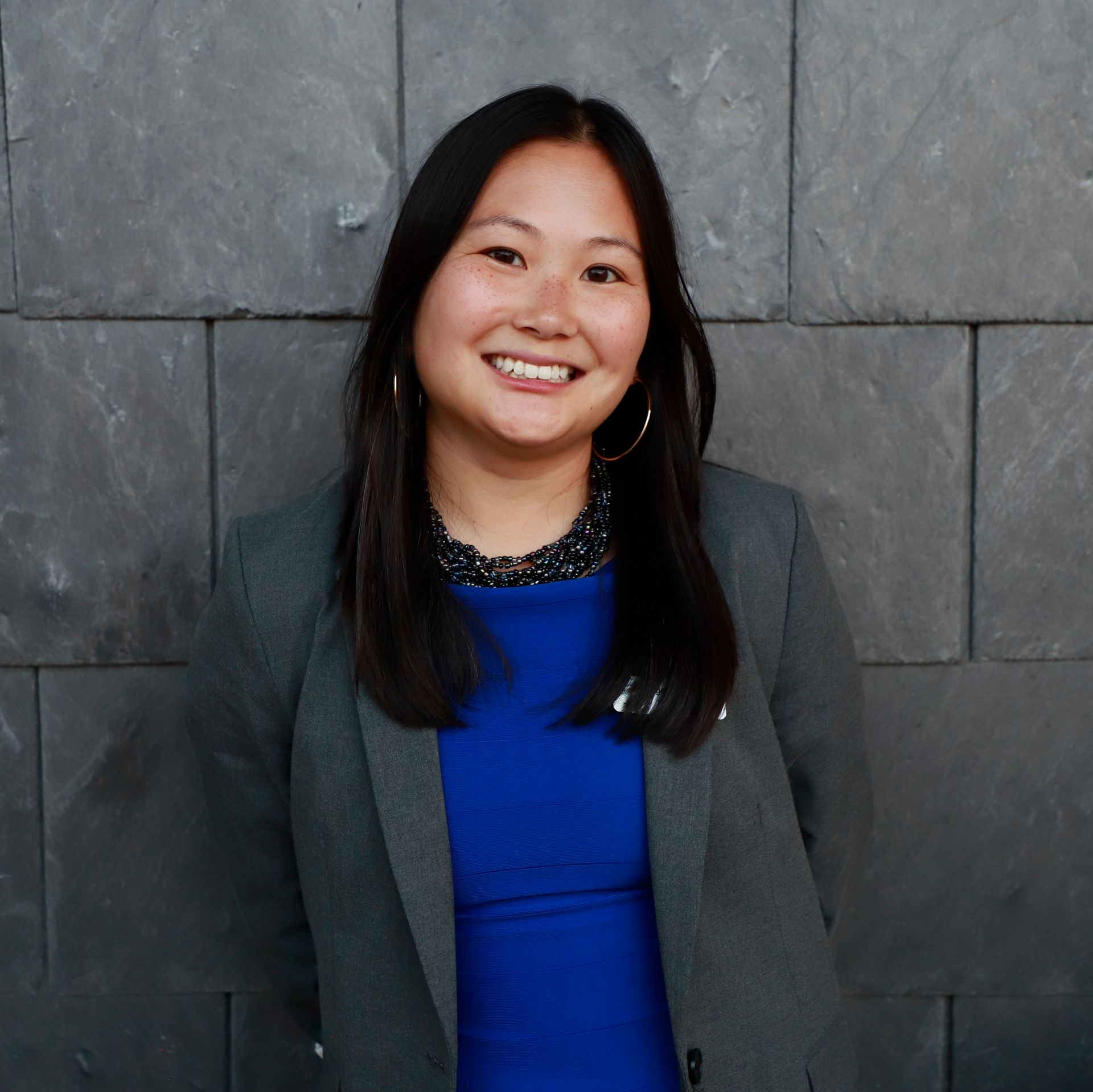 Smiling person in a gray blazer and blue top standing against a gray stone wall