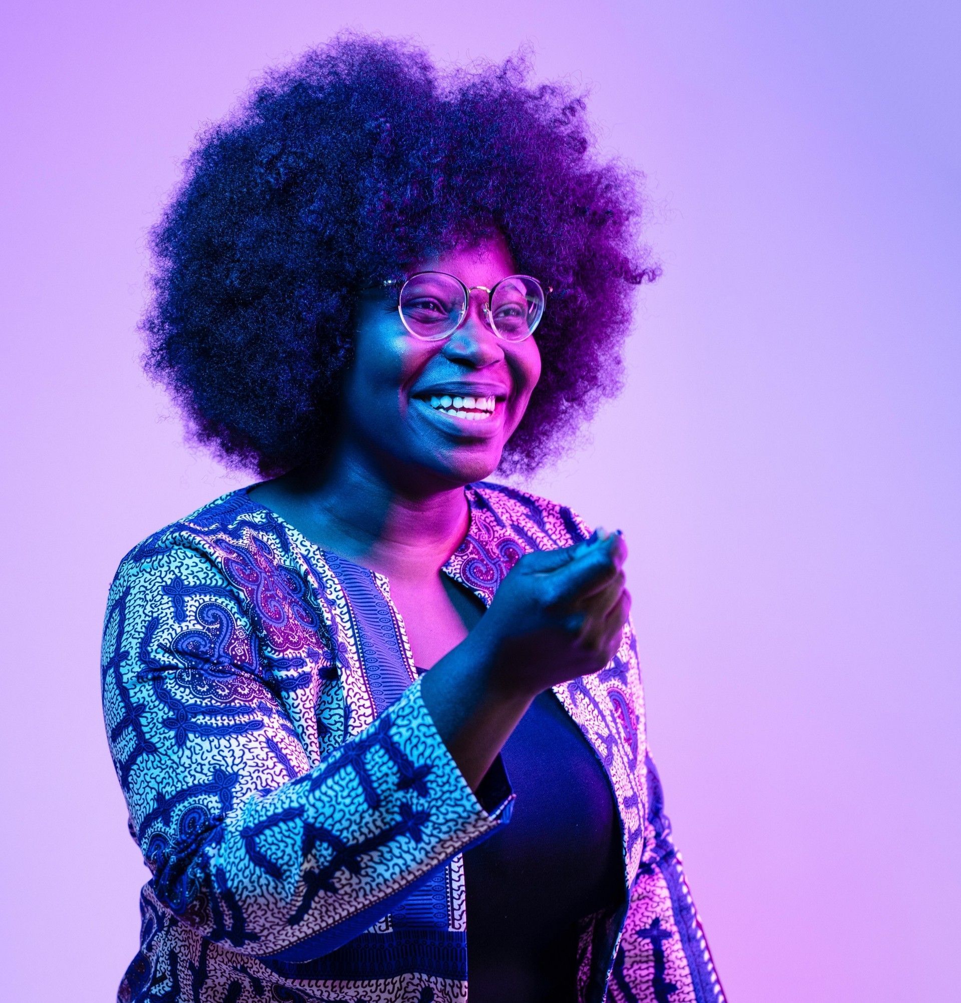 Smiling person in glasses, lit in blue and purple, holding a microphone on stage