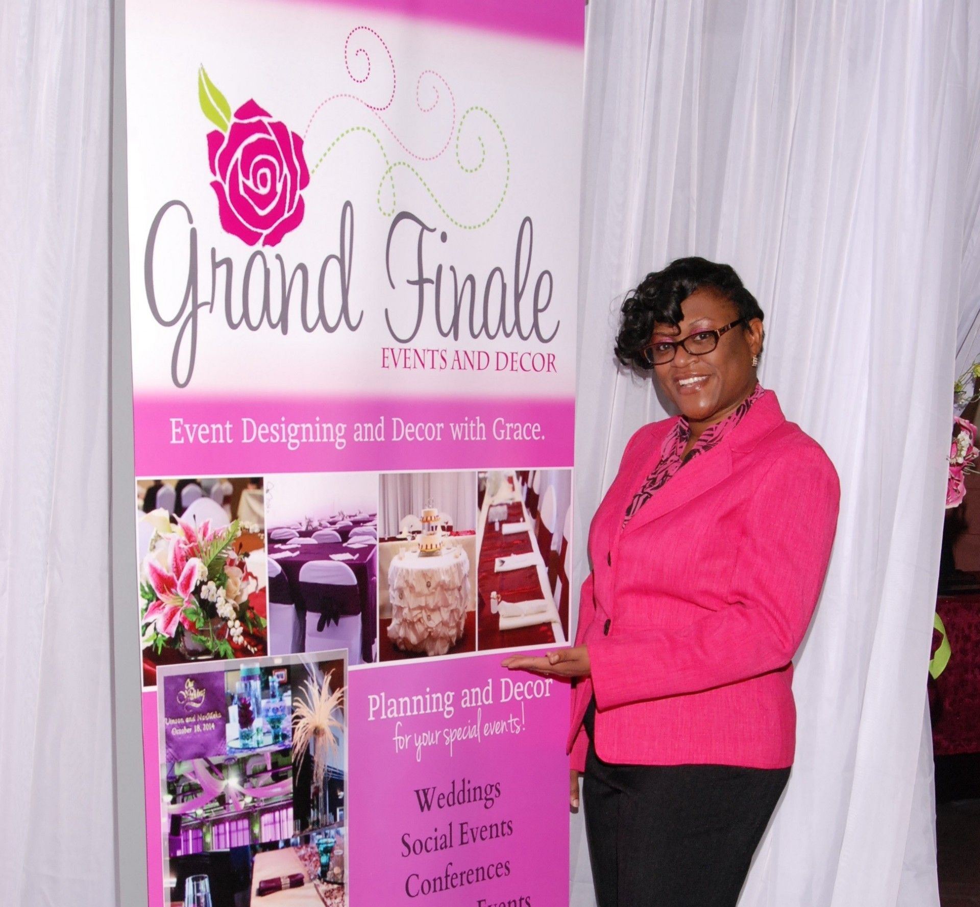 Woman in pink jacket beside Grand Finale event planning display booth with floral decor and photos