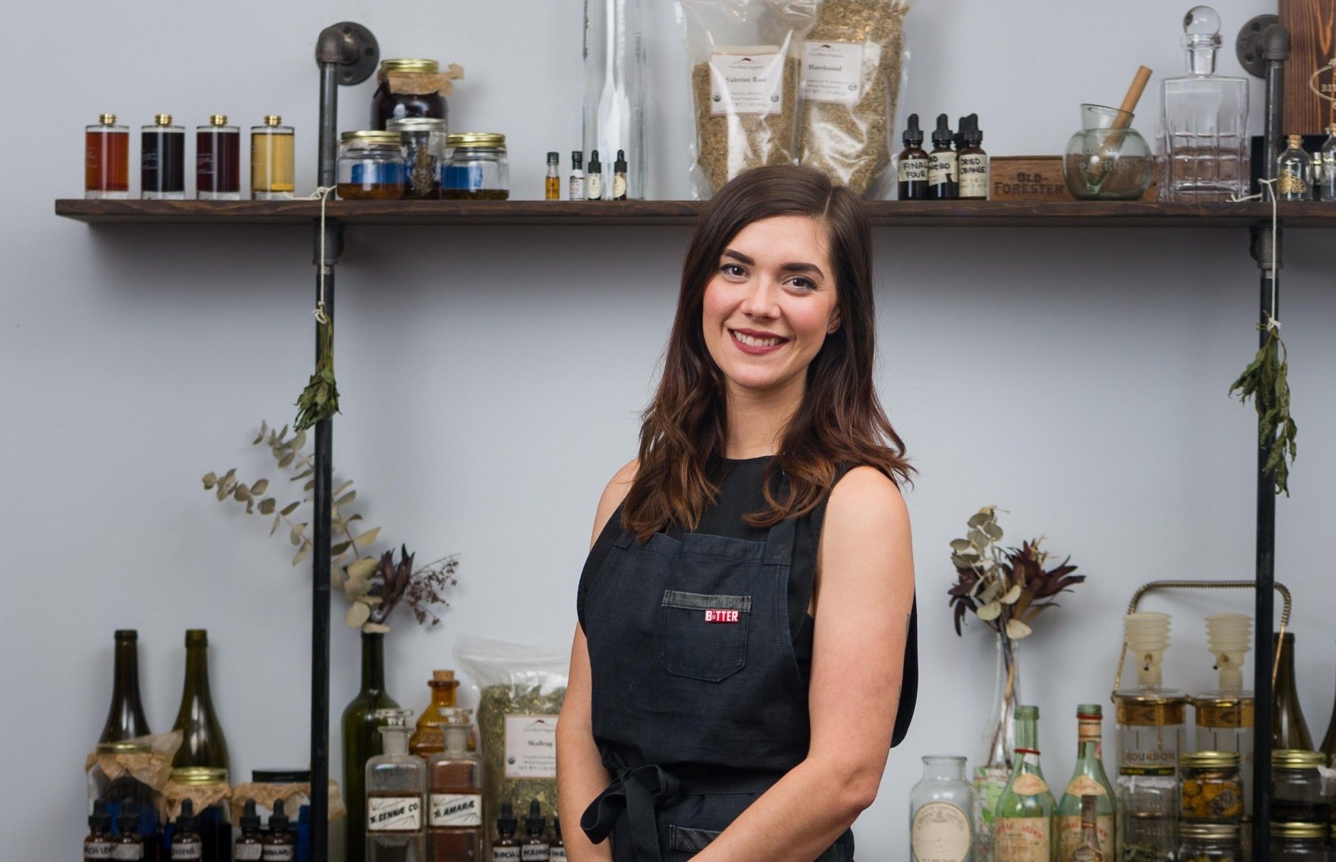 Woman smiling in an apron, standing in a rustic bar with bottles and glassware on shelves.