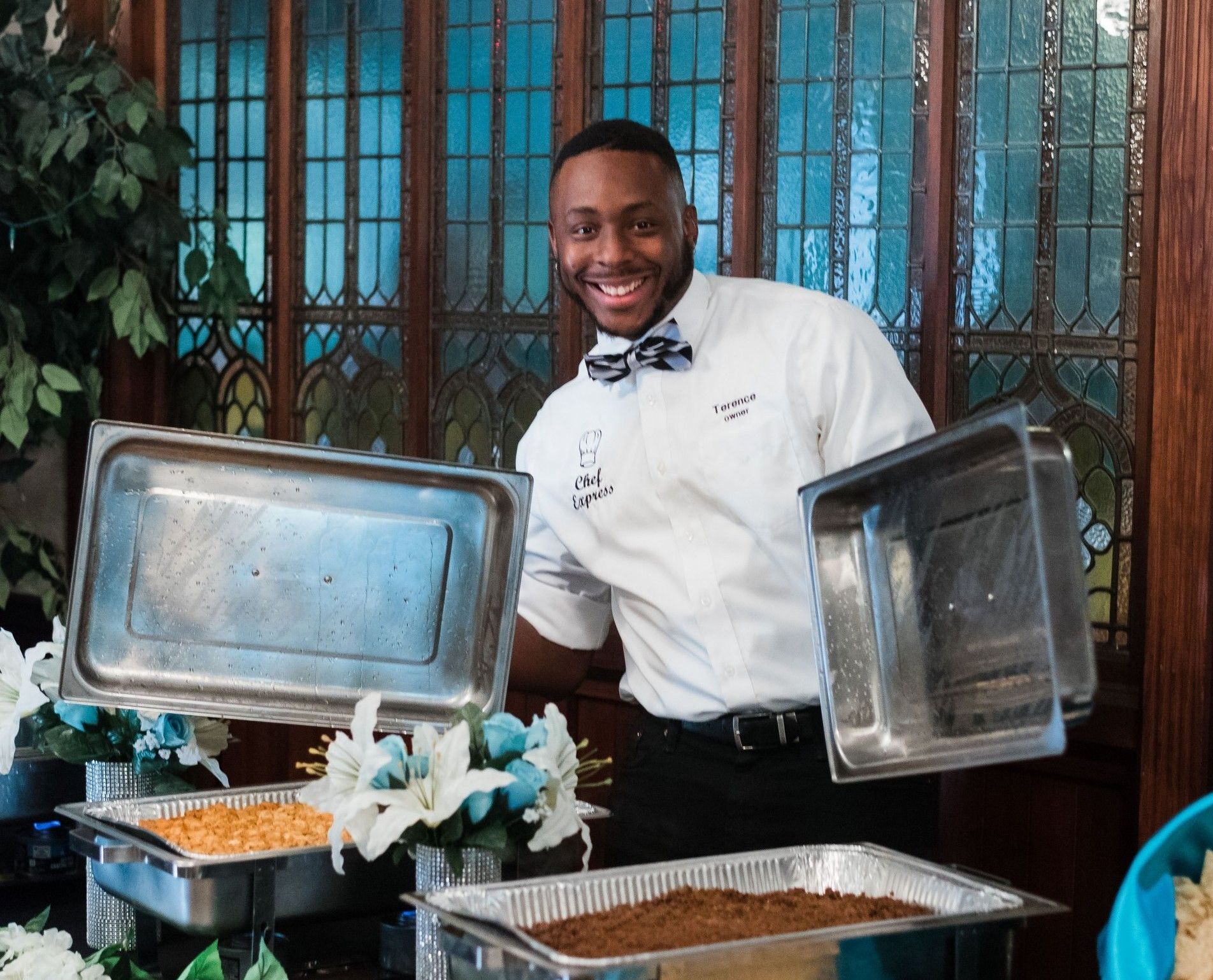 Smiling chef holding open chafing dish lids beside buffet trays in a restaurant setting