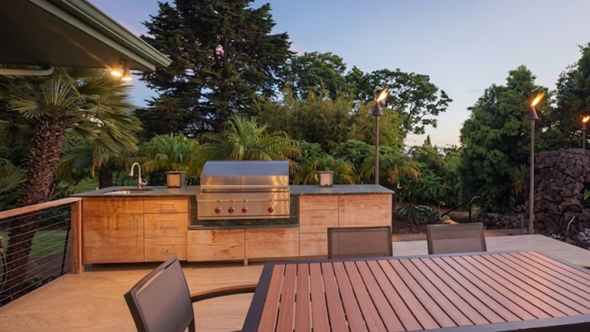 Backyard patio with built-in outdoor kitchen, wooden dining table, and trees at dusk