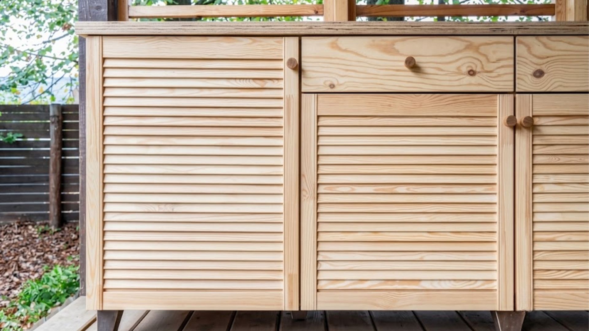 Wooden louvered cabinet doors on a deck, with a wood fence and trees in the background.