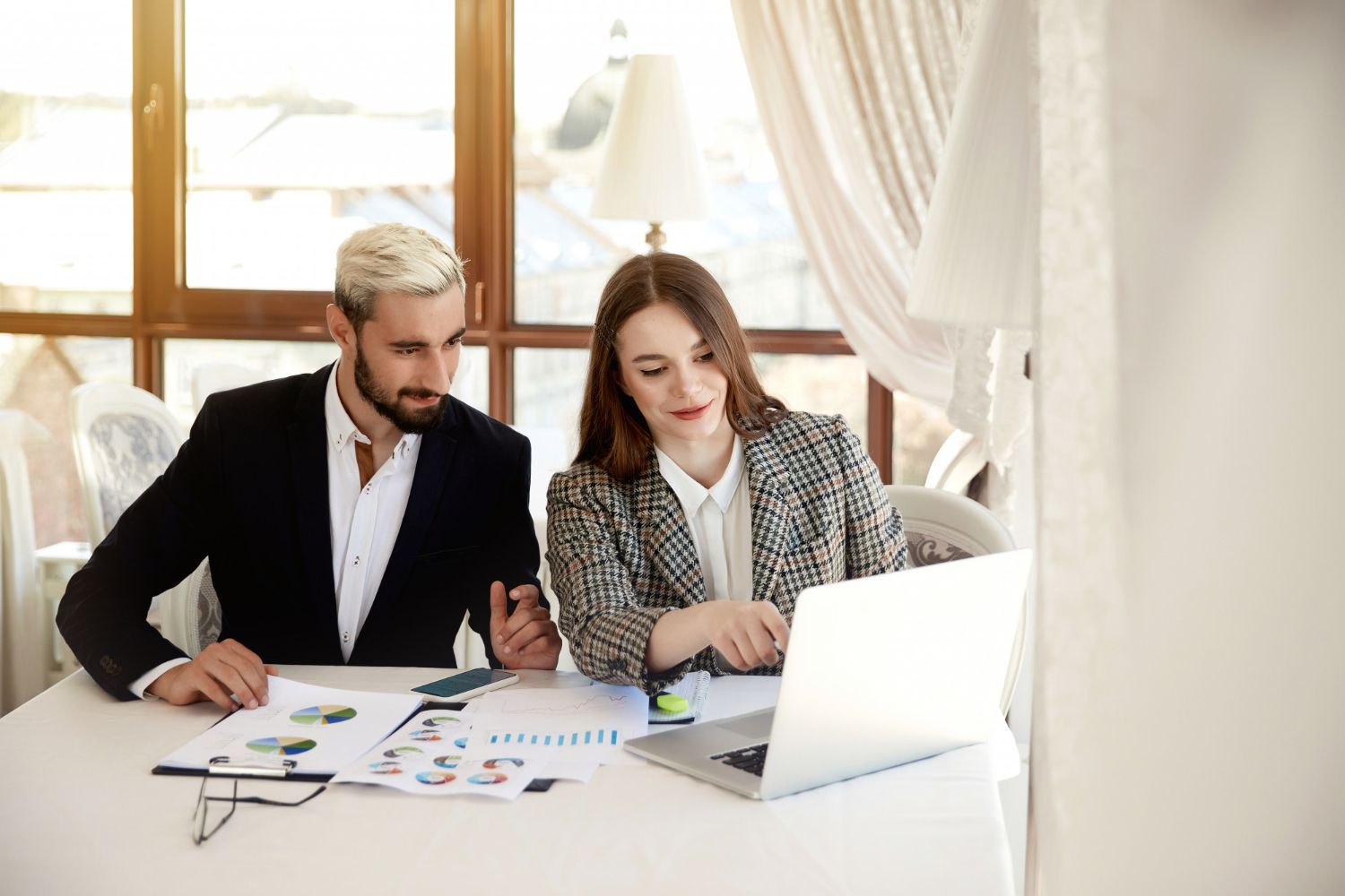Two coworkers review charts at a desk beside a laptop in a bright office