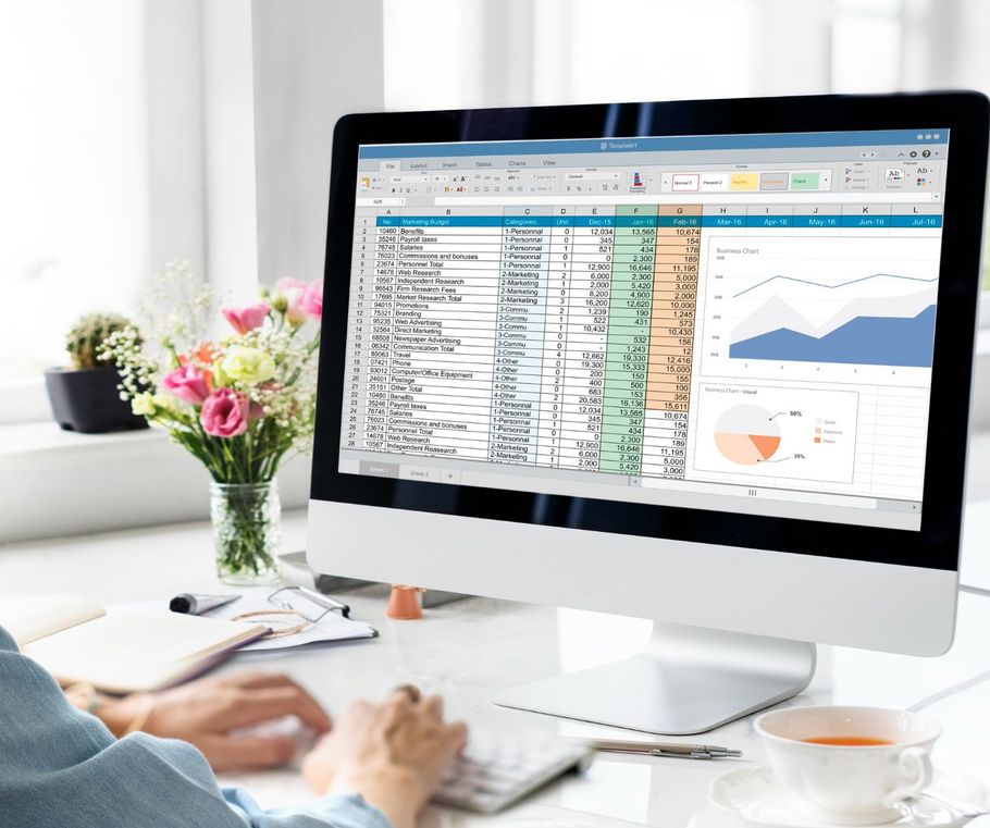 Person working at a desk with a desktop monitor showing charts and a spreadsheet in a bright office