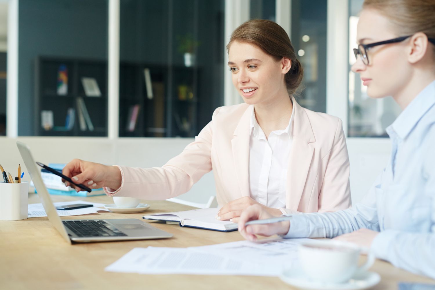 Two coworkers discussing a document at a desk in a bright office, with a laptop open nearby