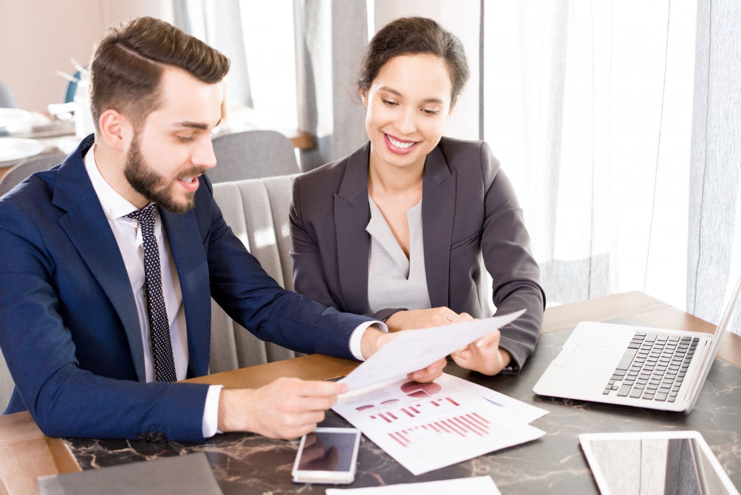 Two coworkers review documents at a desk with a laptop and phone in a bright office.