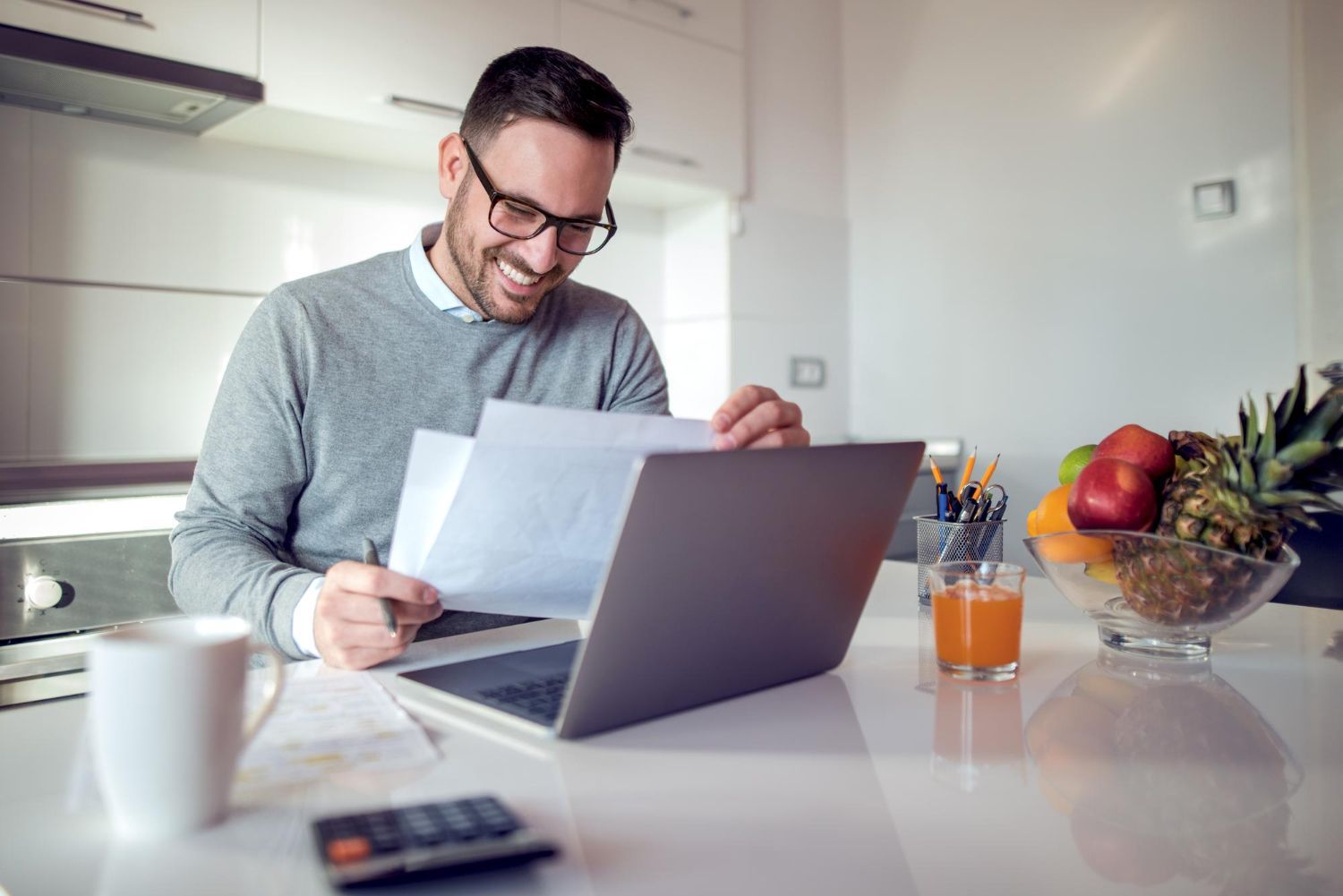 Man reviewing a document beside a laptop at a bright kitchen table with coffee, fruit, and office supplies