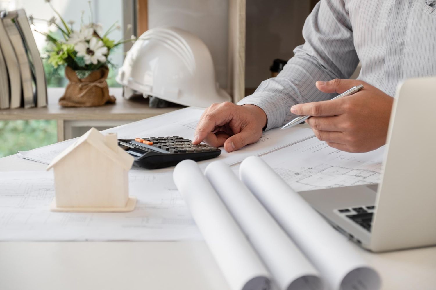 Desk with architectural plans, calculator, laptop, and hard hat; person points at documents.
