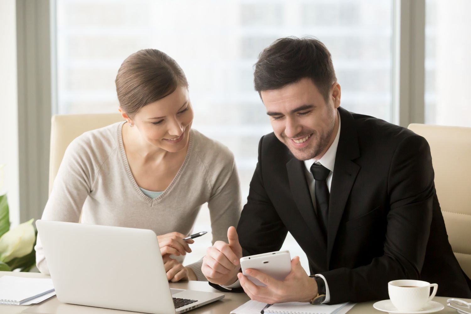 Two professionals reviewing documents at a laptop, smiling in a bright office