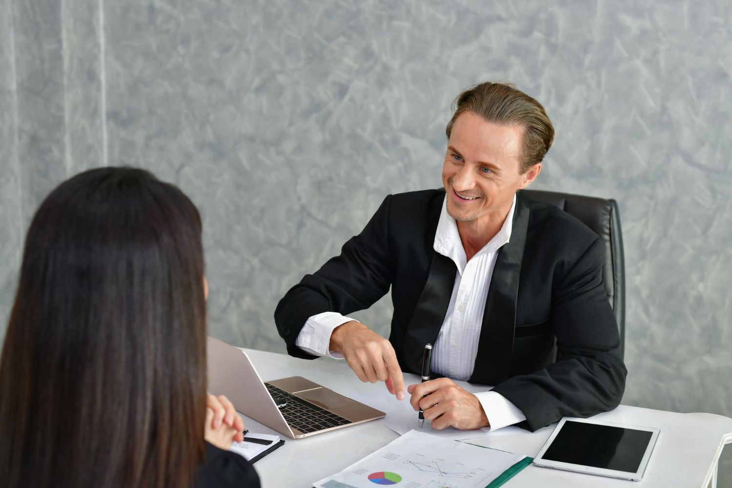Business meeting with two people across a desk, laptop and documents visible, one smiling and gesturing.