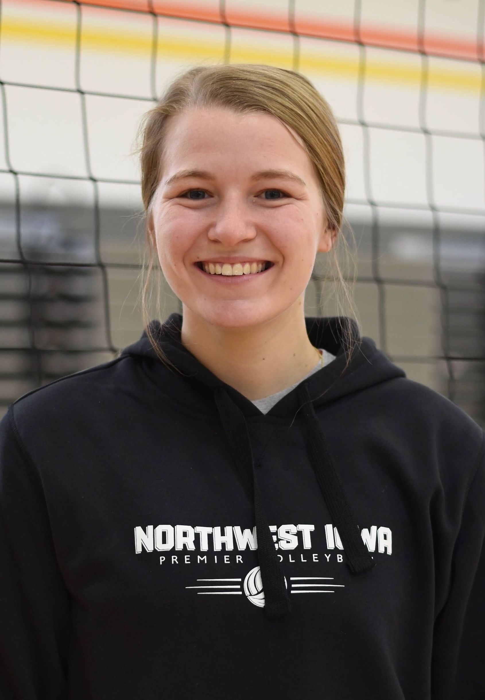 A woman in a black jacket is smiling in front of a volleyball net.