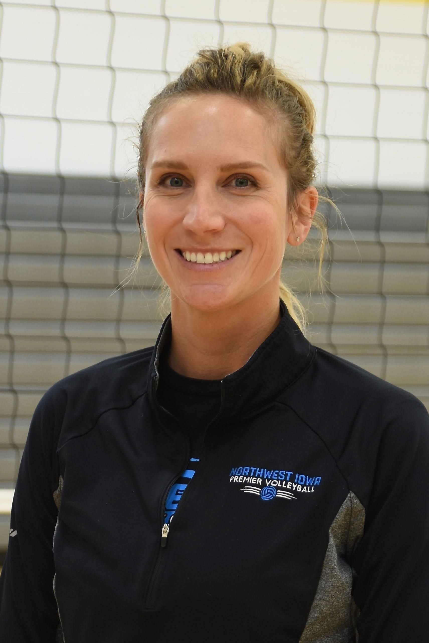 A woman in a black jacket is smiling in front of a volleyball net.
