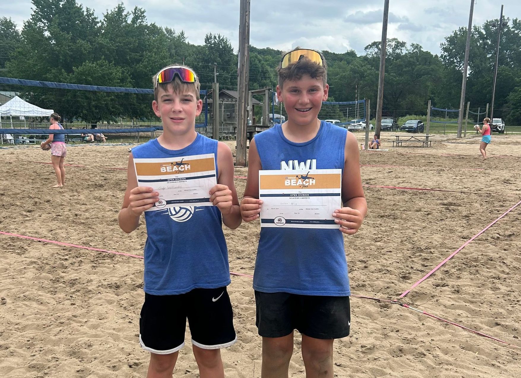 Two young boys are standing on a sandy beach holding certificates.