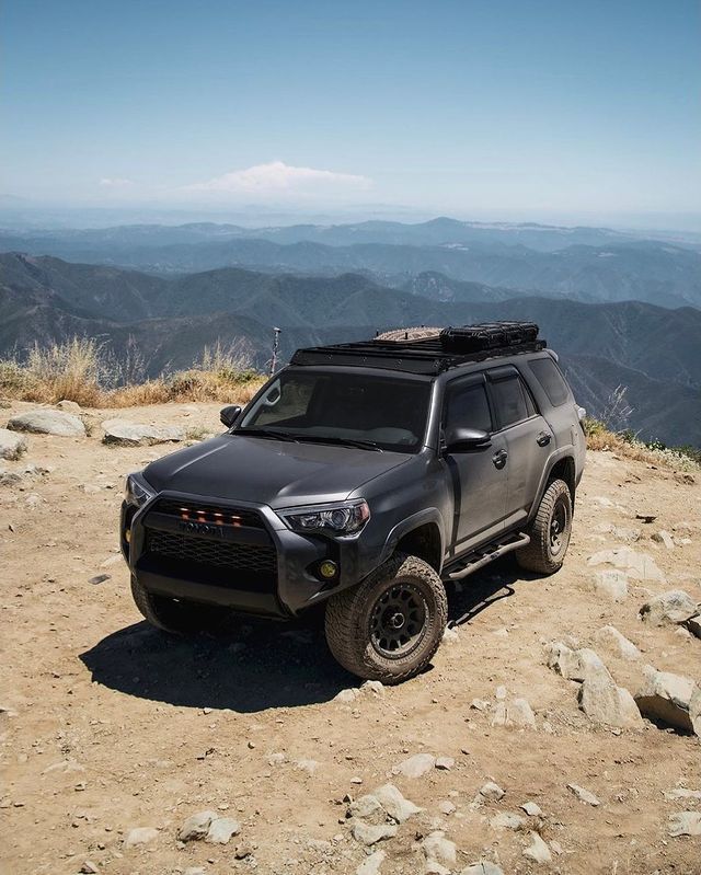 A black suv is parked on top of a dirt hill with mountains in the background.