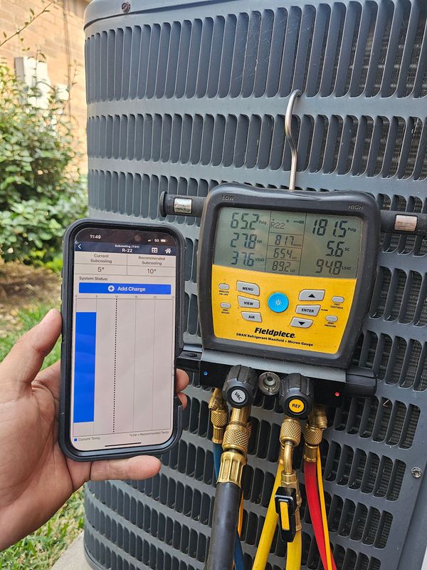 Hand holding a smartphone beside a yellow HVAC manifold gauge connected to outdoor AC unit wires and hoses