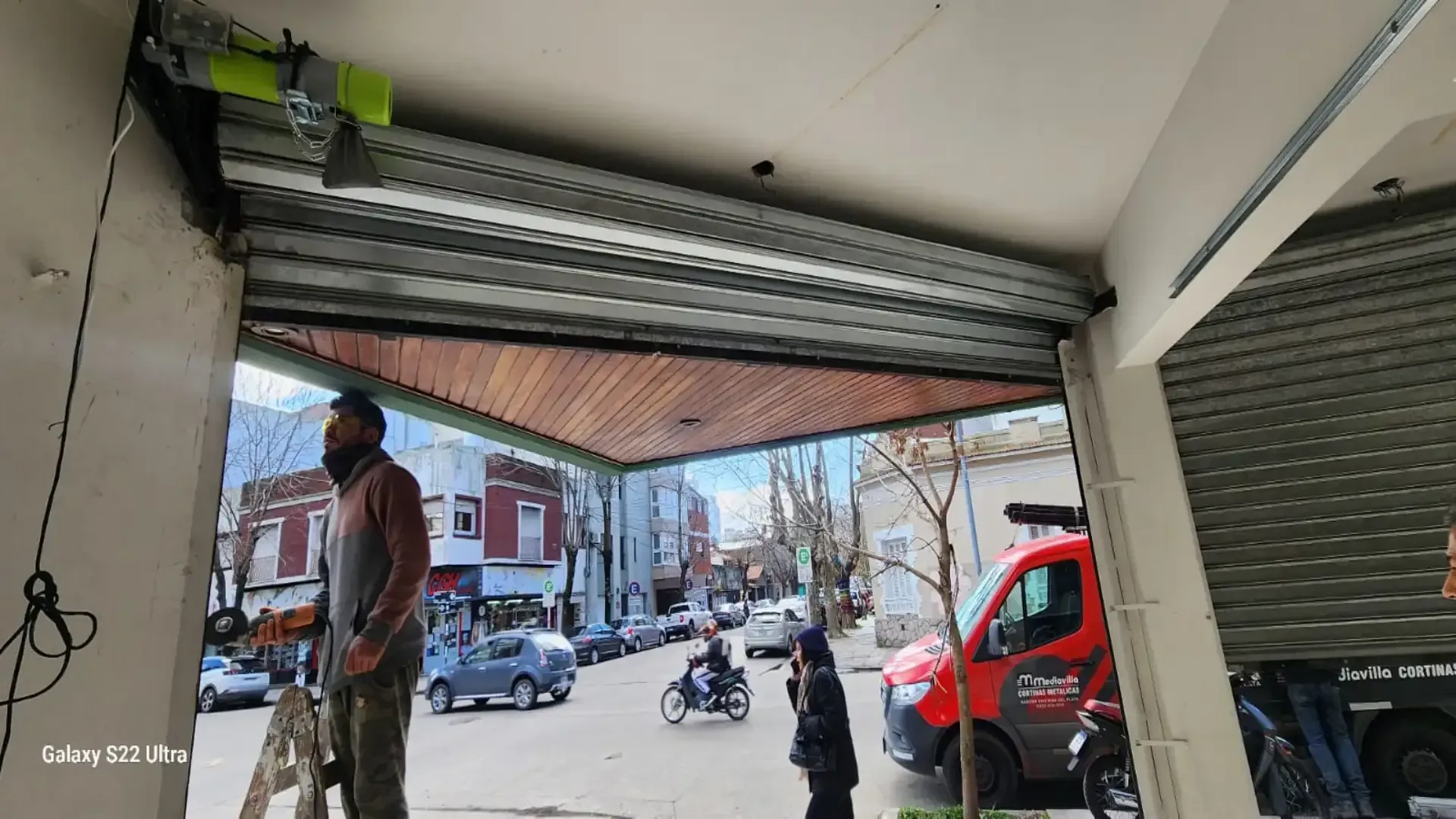Persona trabajando cerca de un local comercial parcialmente abierto con la calle y patinetes aparcados en el exterior.