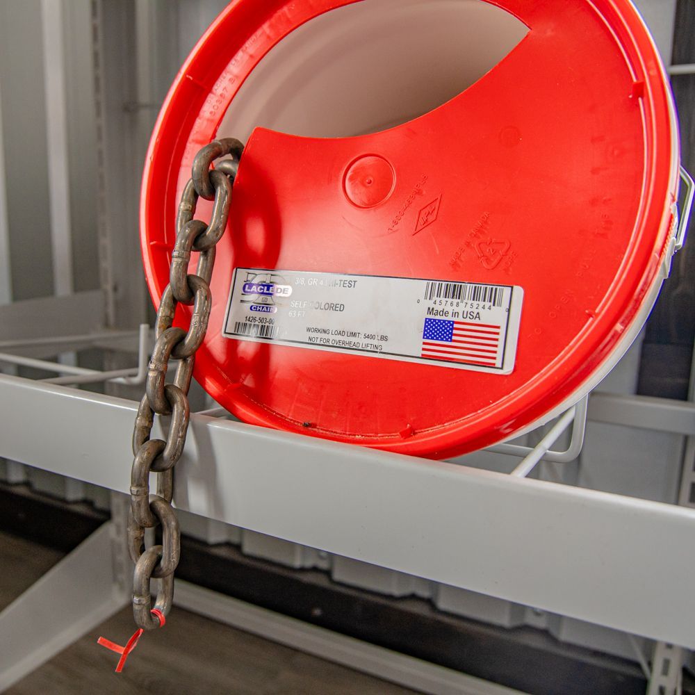 A length of rusty metal chain hangs from the hole of a bright red plastic bucket lid resting on a white metal shelf.