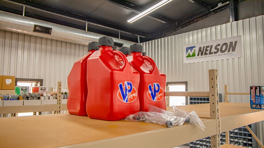Several red VP Racing fuel jugs sit on a wooden shelf in a warehouse with a Nelson sign in the background.