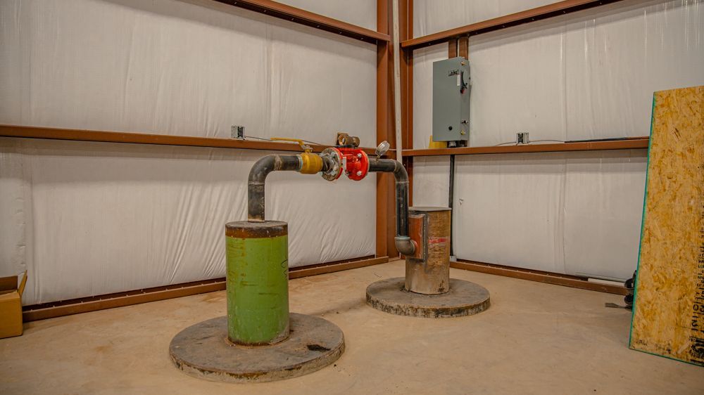 A loop of black pipe connects two vertical cylinders, one green and one rusted metal, on a concrete floor in a workshop.