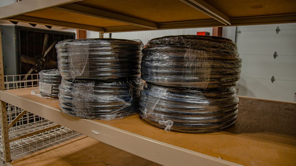 Four large, shrink-wrapped coils of black cable sit on a tan wooden shelf in a warehouse setting.